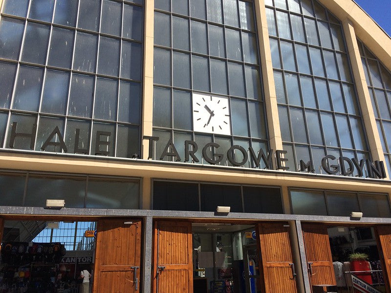 The exterior of a market hall with large glass windows, a clock showing 11:21, and signage that reads "Hale Targowe w Gdyni" above open wooden doors. Sunlight is shining on the building.