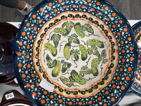 A colorful Polish Pottery ceramic plate with a decorative border of orange dots and black swirls surrounds the central pattern of green leaves and cucumbers on a speckled blue and white background. A small sticker with numbers is visible on the plate.